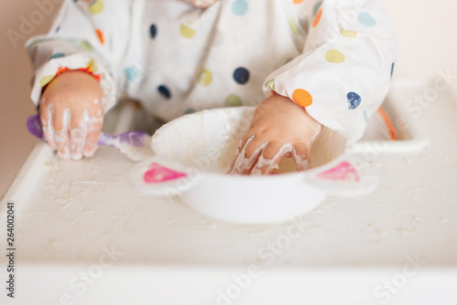 a little girl in a t-shirt and plate sitting in a child's chair eating with hands cereals with yogurt food