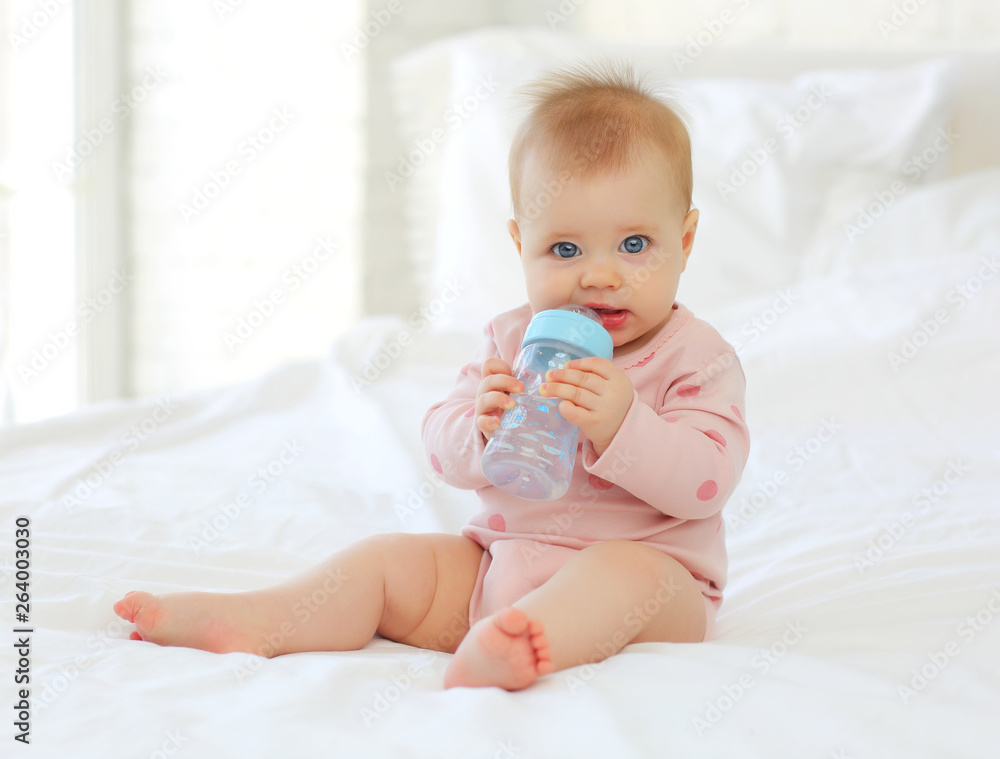 Charming baby 9 months old sitting on the bed in pink clothes with a bottle of water