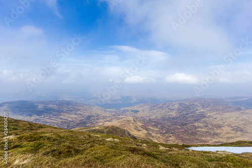 A view of a mountain valley with grass, snow and lake in the background under a majestic blue sky and white clouds © Dolwolfian