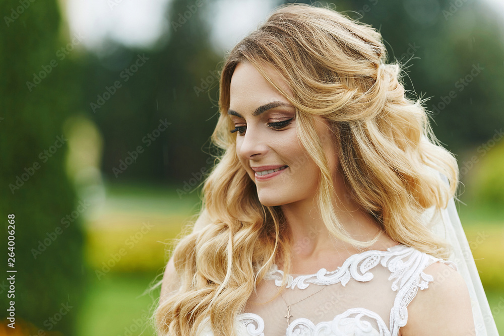 Fototapeta premium Young blonde bride with a beautiful smile and with stylish wedding hairstyle in lace white dress outdoors in the morning of the wedding day