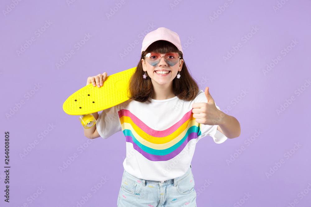 Joyful teen girl in vivid clothes, heart eyeglasses hold yellow skateboard, showing thumb up isolated on violet pastel wall background. People sincere emotions, lifestyle concept. Mock up copy space. Joyful teen girl in vivid clothes, heart eyeglasses hold yellow skateboard, showing thumb up isolated on violet pastel wall background. People sincere emotions, lifestyle concept. Mock up copy space.