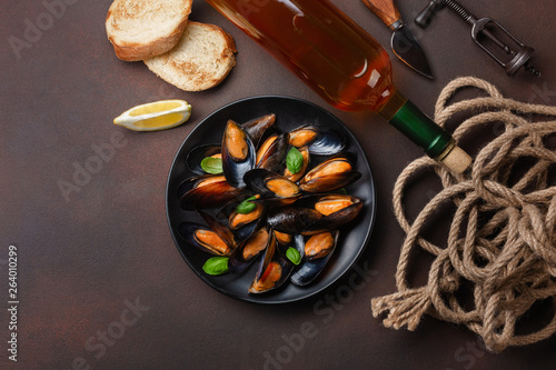 Seafood mussels and basil leaves in a black plate with wine bottle, corkscrew, rope, bread slice, fork and knife on rusty background