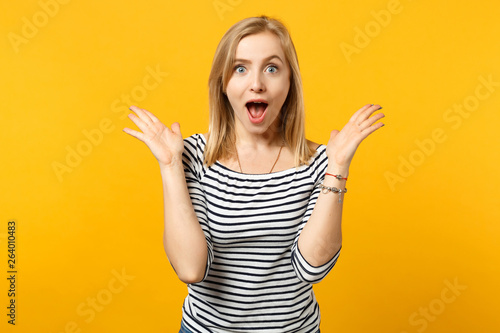 Excited young woman in striped clothes keeping mouth wide open, spreading hands isolated on yellow orange wall background in studio. People sincere emotions, lifestyle concept. Mock up copy space.