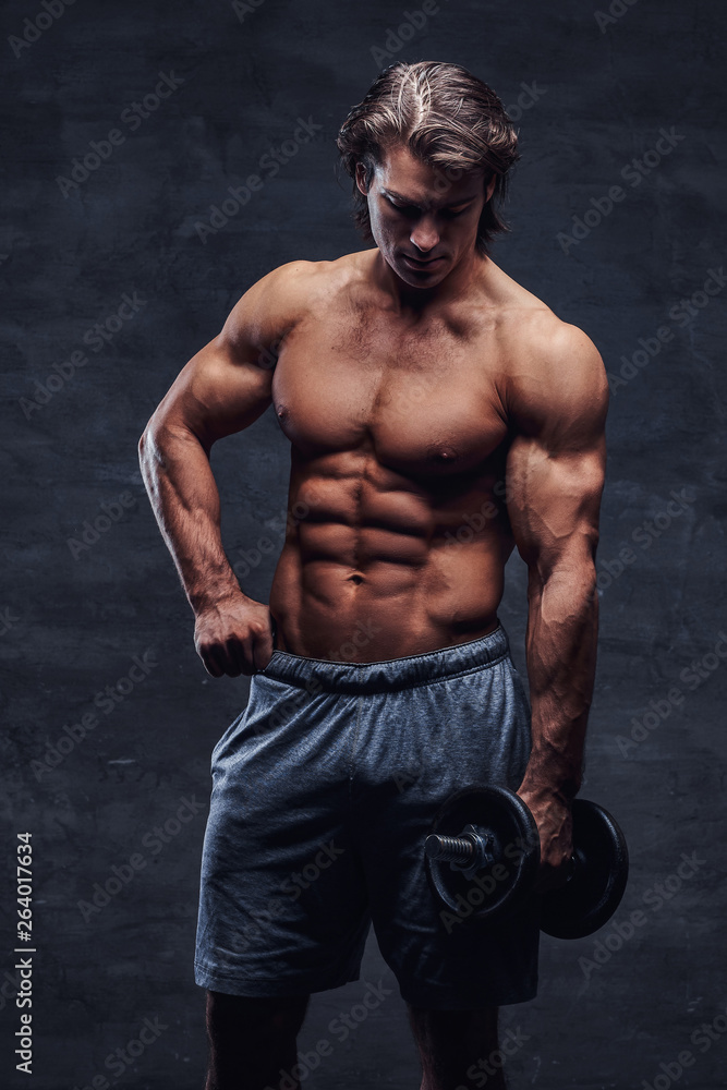 Attractive shirtless bodybuilder is doing exercise with dumbbell. There is dark background.