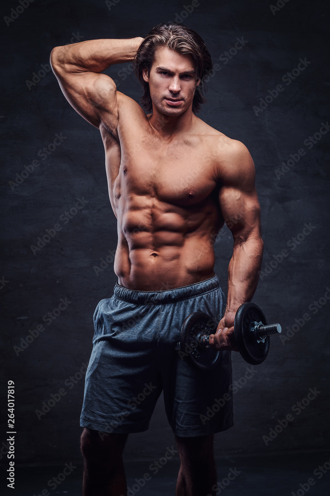 Attractive shirtless bodybuilder is posing with dumbbell. There is dark background.