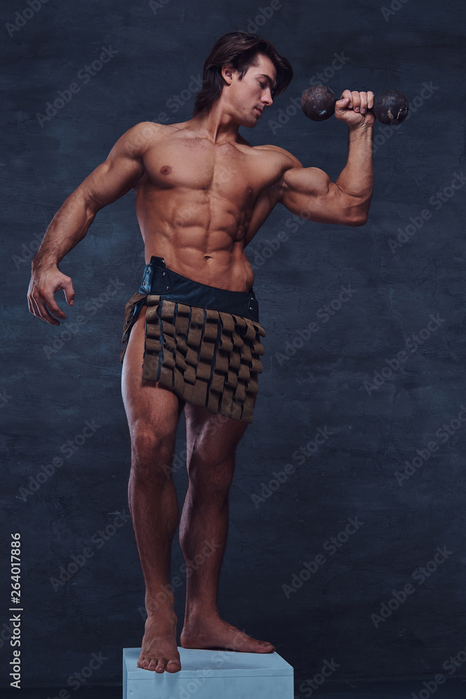 Obraz premium Beautiful strong bodybuilder is posing on white pedestal with naked torso, dumbbell and gladiator's bandage.
