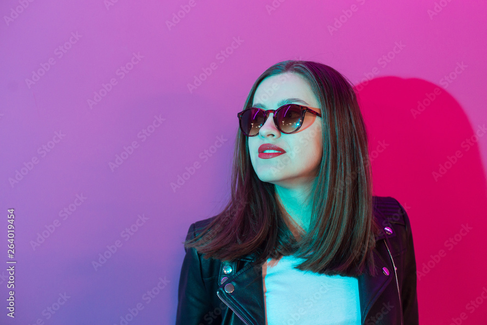 Fashioned girl in leather jacket in dark glasses in pink anf blue neon lights background. Fashioned girl in leather jacket in dark glasses in pink anf blue neon lights background.