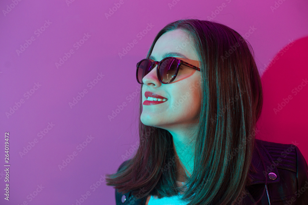 Obraz premium Fashioned girl in leather jacket in dark glasses in pink anf blue neon lights background.