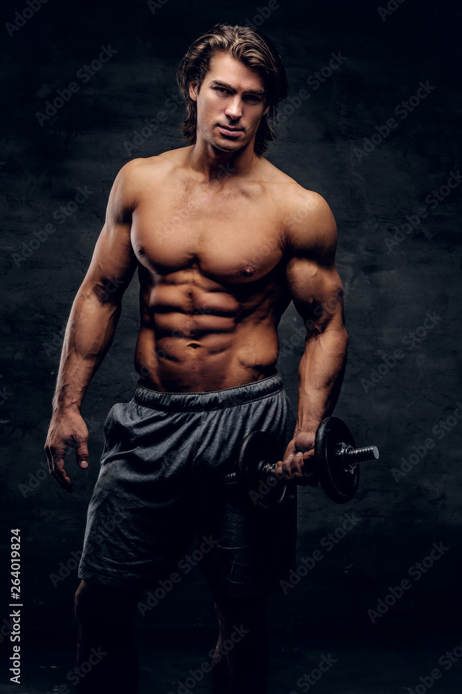 Beautiful young bodybuilder is posing with dumbbell. He has naked torso.