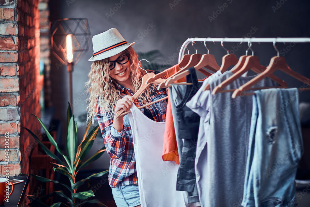 Happy young woman in hat and glasses is getting ready for summer at modern loft. She wear checkered shirt. Woman is standing near clothes rack.