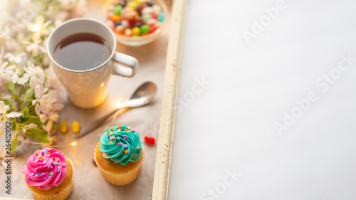 Bright cakes. Cupcakes with a cup of coffee or tea and color dots on a tray with a flowering branch and luminous garland on a white background
