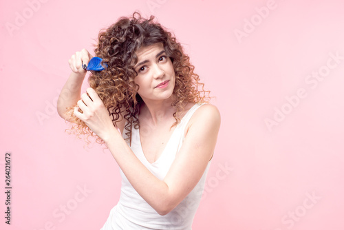 Beautiful woman with a smile on her face with curly hair in a white T-shirt combing her blue curls with a blue comb isolate pink background.