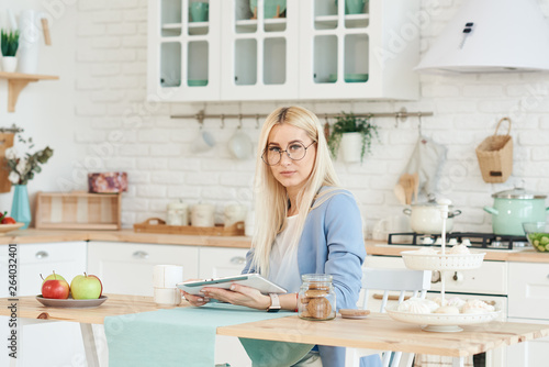 Freelance concept. Beautiful business woman in casual clothes and glasses is examining documents and smiling while working with a laptop in kitchen. Working at home