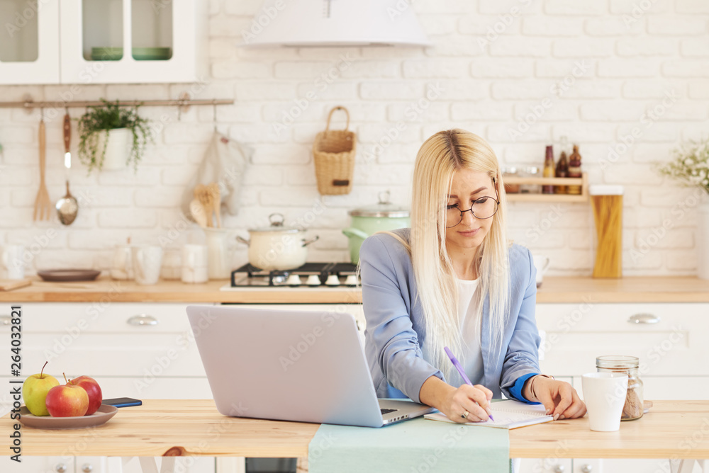Freelance concept. Beautiful business woman in casual clothes and glasses is examining documents and smiling while working with a laptop in kitchen. Working at home Freelance concept. Beautiful business woman in casual clothes and glasses is examining documents and smiling while working with a laptop in kitchen. Working at home