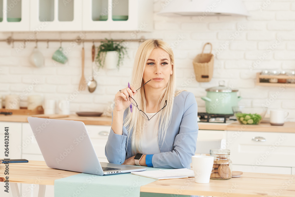 Freelance concept. Beautiful business woman in casual clothes and glasses is examining documents and smiling while working with a laptop in kitchen. Working at home Freelance concept. Beautiful business woman in casual clothes and glasses is examining documents and smiling while working with a laptop in kitchen. Working at home