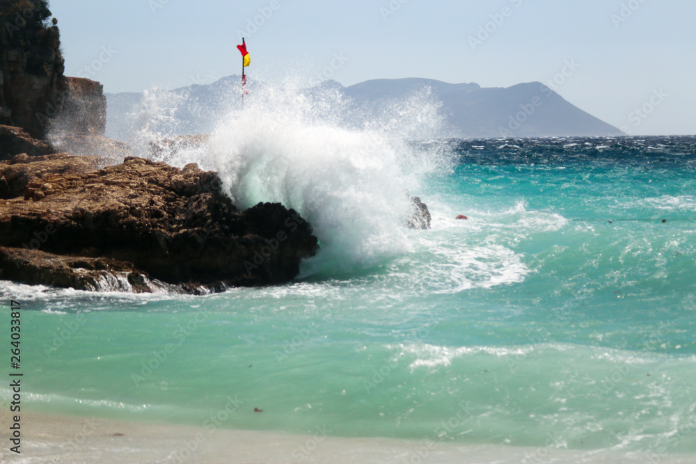 Fototapeta premium Waves coming from the sea are blasting on the rocks. Water color is turquoise and a red yellow flag is on the rock. Windy and stormy weather at Mediterranean Sea. Buyukcakil Beach, Antalya Turkey