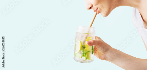 Close up of a woman's hand holding a glass of tropical mojito cocktail and sipping it through a straw on cyan pastel background with copy space. Summer time vacation concept.