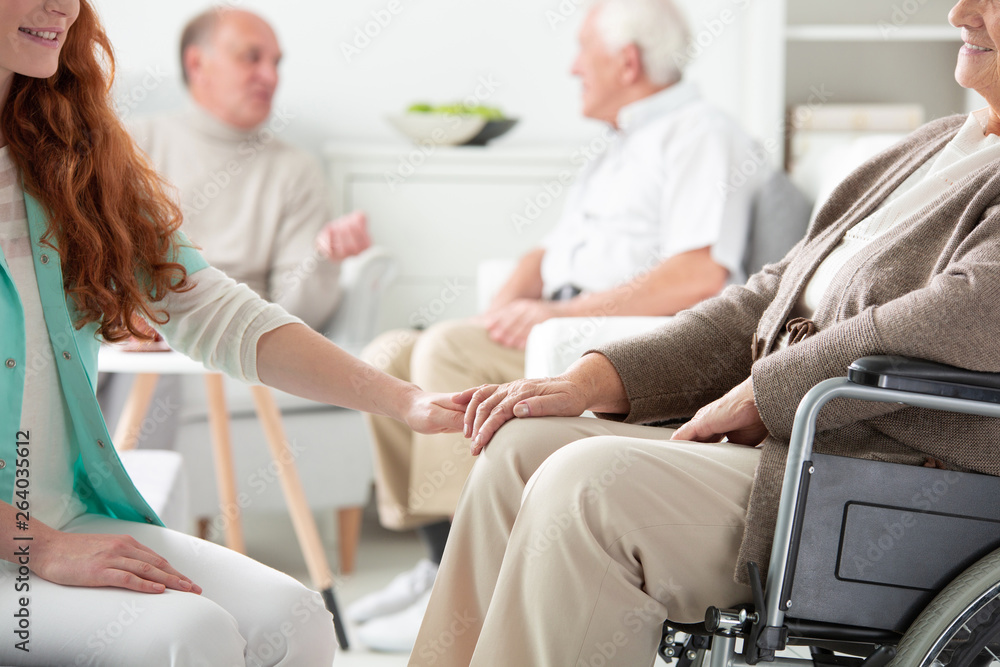 Close-up on friendly nurse holding hand of smiling senior person in the wheelchair
