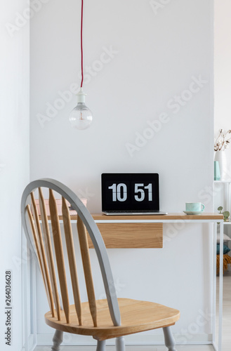 Clock screensaver on a laptop in bright home office interior with wooden chair, simple desk and bare light-bulb. Real photo