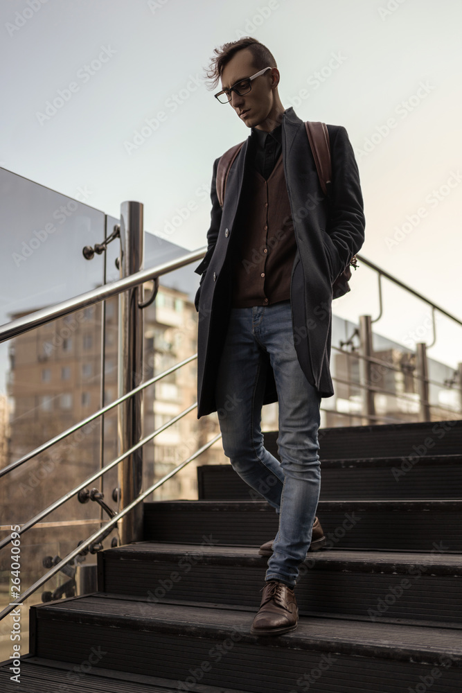 Young stylish man coming down the stairs. Student wearing in black coat, jeans, leather boots, glasses and backpack.