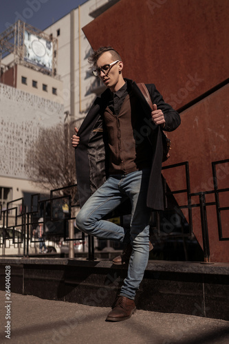 Man street style. Casual clothes. Young student wearing in coat, jeans, boots,backpack, glasses.