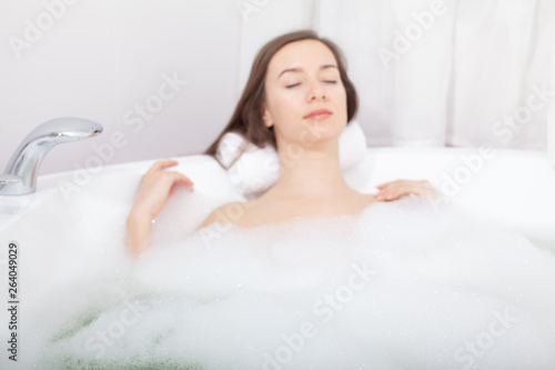 Young pretty woman relaxes in bathtube with soap foam. Spa procedures in bathroom at home or hotelroom. Skin care and moisturizing for youth preservation.