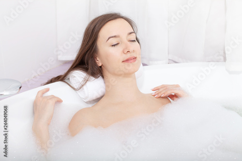 Young pretty woman relaxes in bathtube with soap foam. Spa procedures in bathroom at home or hotelroom. Skin care and moisturizing for youth preservation.
