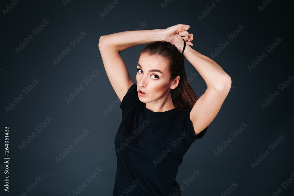 sporty-looking girl in a black T-shirt and leggings on a dark background tying a tail on her head sporty-looking girl in a black T-shirt and leggings on a dark background tying a tail on her head