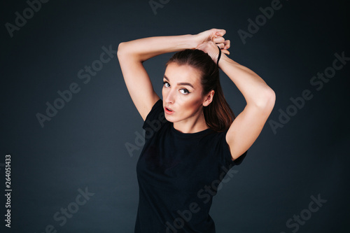 sporty-looking girl in a black T-shirt and leggings on a dark background tying a tail on her head