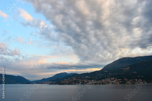 A bay seen at sunset. Grey, pink and white clouds stretch across a blue sky. Rocky hills with mountains in the background surround the bay. © Anne