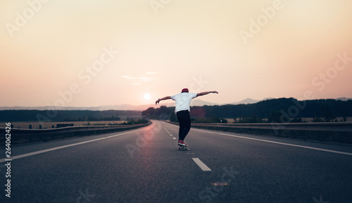 Man with arms outstretched riding a skateboard on the motorway road toward the setting sun in the background. The background is slightly blurred, focus on a skateboarder in the foreground. © Zdena Venclik
