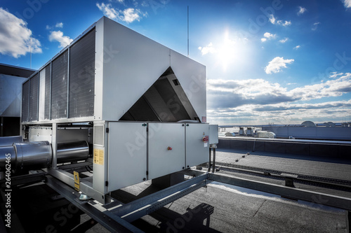 A huge air conditioning unit on the roof of the building. In the background of blue sky with shining sun. Focus is at the front of the air conditioner, the other parts of image slightly blurred.
