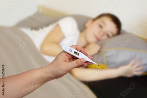 female hand holds white thermometer on a blurred background with a lying boy, light background