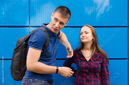 guy in blue T-shirt and backpack on his back next to girl dressed in blue jeans and plaid shirt against blue wall