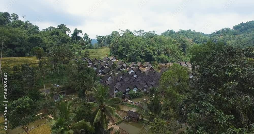 Aerial drone view above lush tropical green tree tops overlooking tranquil Kampung Naga remote rural village rooftops & lush colourful scenery. Aerial drone view above lush tropical green tree tops overlooking tranquil Kampung Naga remote rural village rooftops & lush colourful scenery.