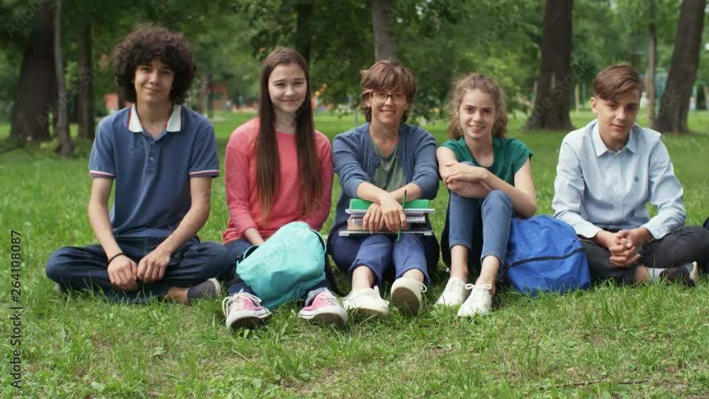 Zoom in of classmates and their female middle-aged teacher sitting on lawn, looking at camera and smiling Zoom in of classmates and their female middle-aged teacher sitting on lawn, looking at camera and smiling