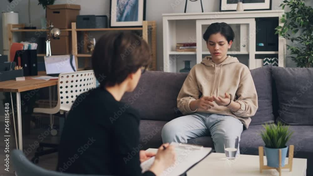 Sad lonely teenager is opening up to professional therapist in psychologist's office in clinic. Upset boy is sitting on couch gesturing and telling story. Sad lonely teenager is opening up to professional therapist in psychologist's office in clinic. Upset boy is sitting on couch gesturing and telling story.