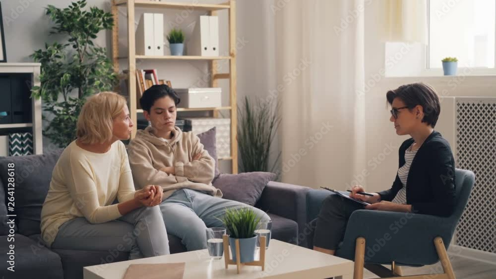 Worried parent mature woman is discussing parenting problems with therapist while teenage boy is sitting with crossed arms feeling unhappy. Family and psychology concept. Worried parent mature woman is discussing parenting problems with therapist while teenage boy is sitting with crossed arms feeling unhappy. Family and psychology concept.