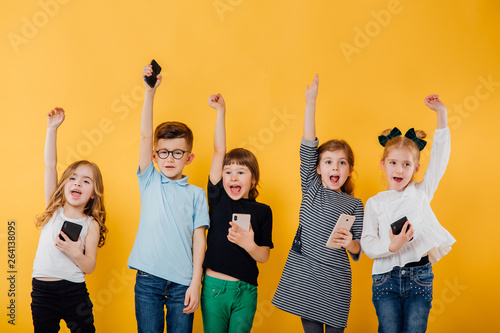 5 beautiful children use the smartphone, raise their hands up, they are happy, isolated yellow background, copy space photo