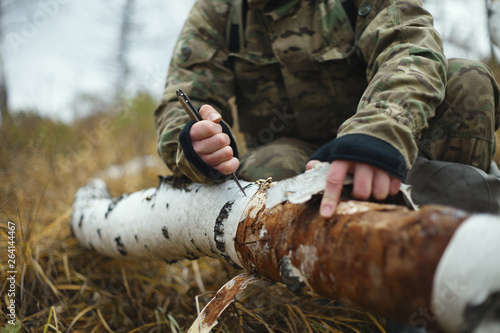 Man traveler in camouflage removes bark from a fallen birch trunk for kindling fire closeup. Extreme travel. Survival in the wild. photo