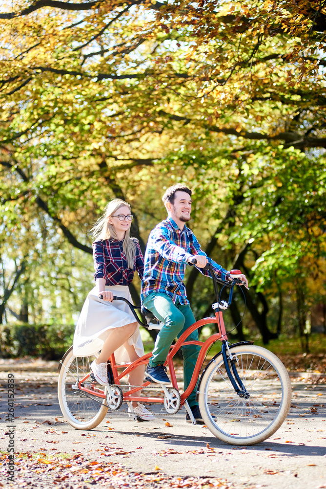 Fototapeta premium Young tourist pair, handsome bearded man and attractive blond long-haired woman cycling together tandem double bike by sunny alley with golden leaves on tall trees background in the fall