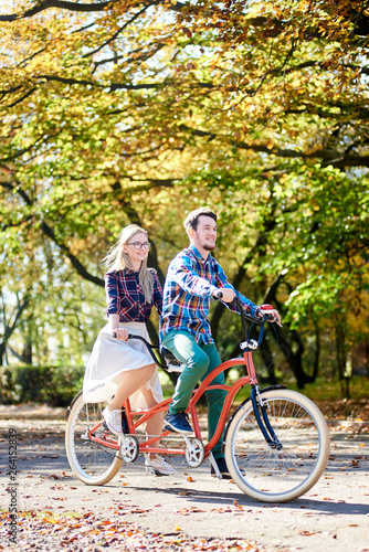 Young tourist pair, handsome bearded man and attractive blond long-haired woman cycling together tandem double bike by sunny alley with golden leaves on tall trees background in the fall