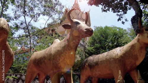 Golden magical animals including a bull, rooster, and parrot guard a Buddhist temple in the forest of Southeast Asia. Camera is low aerial shot (drone), and flies right to reveal more animals and stup photo