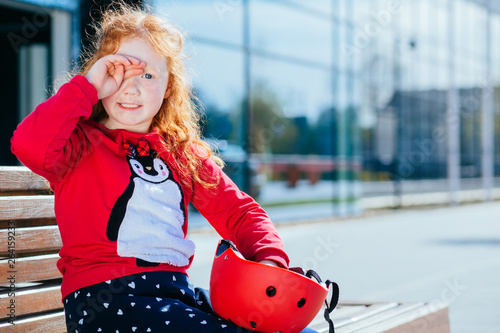 Portrait of a cute curly red hair girl in red shirt rub her eyes, sit on bench, hold orange helmet outdoor over business center on backgriund. Spring, acitivities, childhood, leisure people concept photo