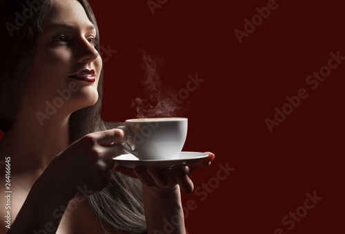 Attractive lady brunette with pleasure holding a coffee cup with light smoke in hand in studio at the bordo background