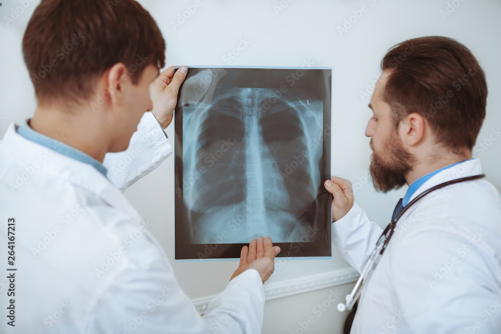 Obraz premium Rear view shot of a two male doctors examining x-ray scan of a patient. Medical team discussing lungs x-ray of a patient. Experienced therapists working at the clinic. Medical service concept