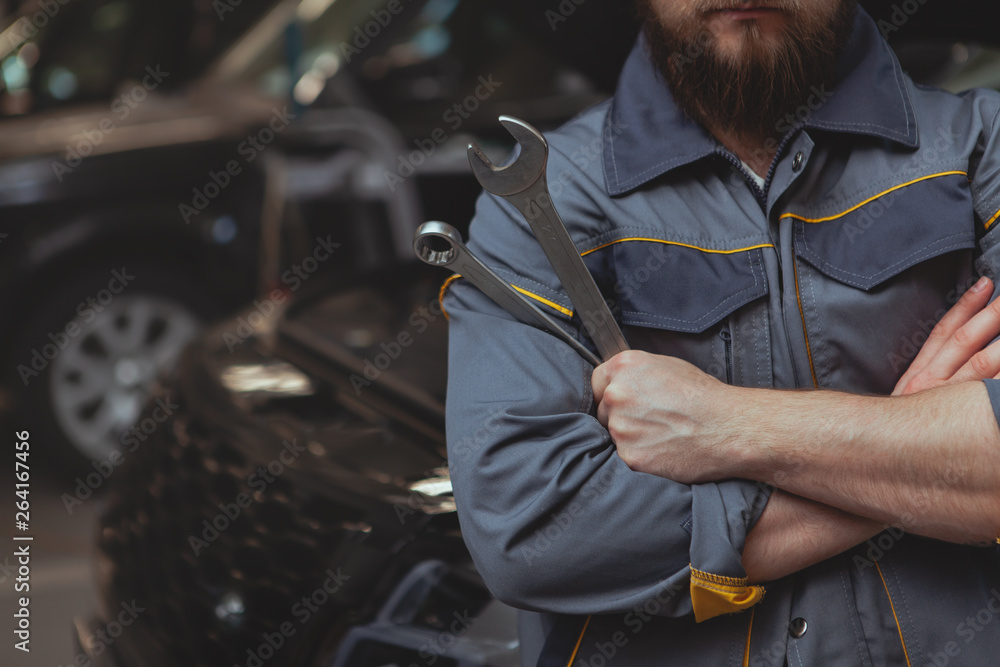 Obraz premium Cropped close up of a bearded car mechanic holding a wrench tool, standing in front of a carat the garage. Unrecognizable automobile technician man at auto workshop, copy space