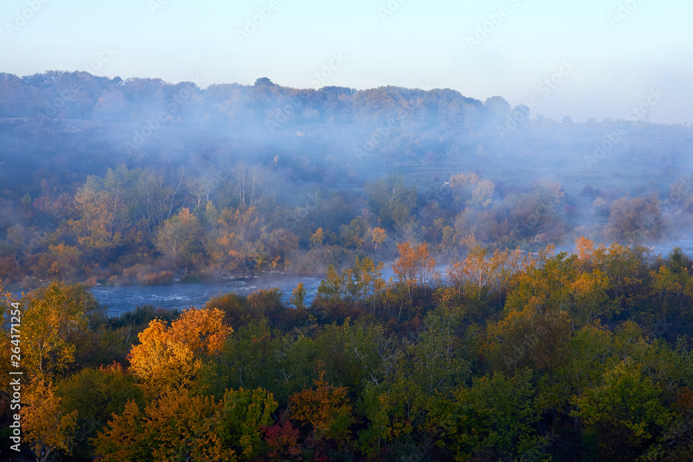 Mist over the river on autumn morning and forest on its bank with yellow colors of the leaves lit by the rising sun. A soft autumn morning, mist over the trees and cold and calm water Mist over the river on autumn morning and forest on its bank with yellow colors of the leaves lit by the rising sun. A soft autumn morning, mist over the trees and cold and calm water