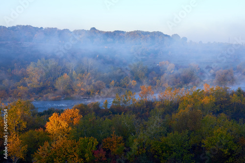 Mist over the river on autumn morning and forest on its bank with yellow colors of the leaves lit by the rising sun. A soft autumn morning, mist over the trees and cold and calm water