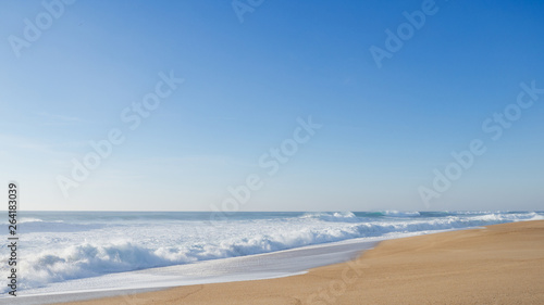 Empty wide coastline with yellow sand in Nazare; huge curving waves crushing on the beach; sunny evening with cloudless sky; clear skyline; sea foam all the way out to the horizon, Portugal